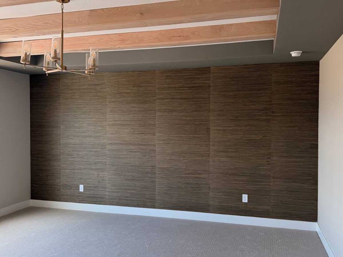 Grasscloth wallpaper installation in Gunnison, Colorado featuring rich dark brown natural texture on a large accent wall beneath exposed wood ceiling beams.