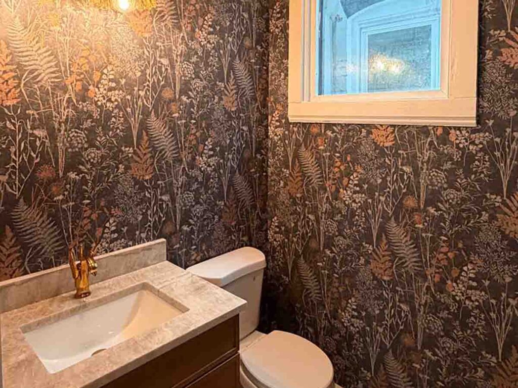 Powder room wallpaper installation in Louisville, Colorado featuring a dramatic dark botanical wallcovering with gold accents, professionally installed around a window and vanity.
