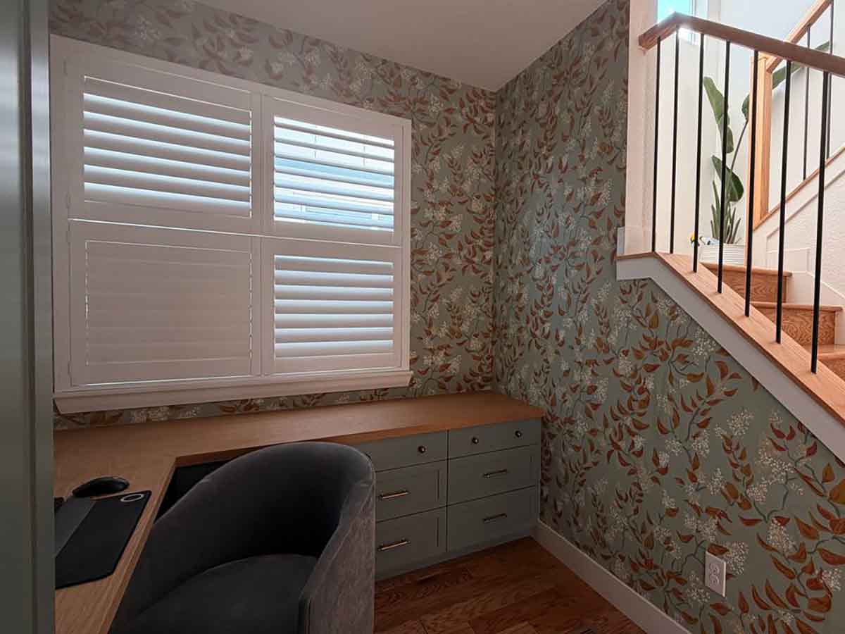 Wallpaper installation in a Pueblo, Colorado home office nook featuring a soft botanical pattern around custom built-in cabinetry, a window with plantation shutters, and a staircase.